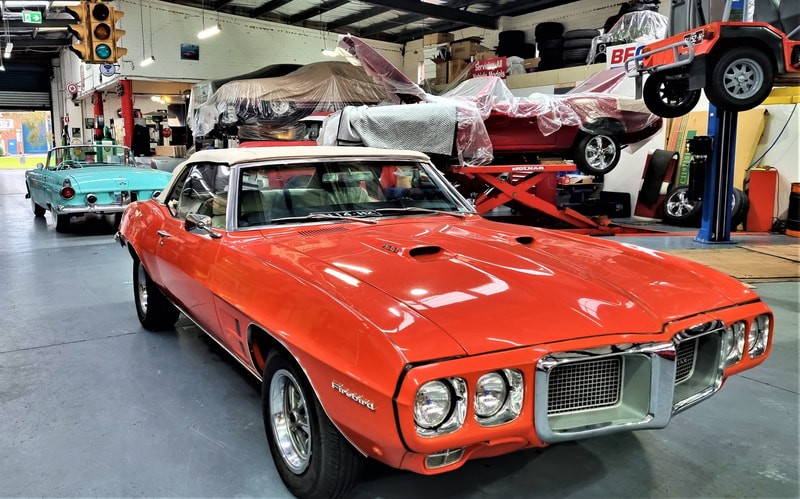 A pair of imported muscle cars in the TGS Muscle Cars garage. One is a bright orange convertible with a white roof, and the other is a bright blue convertible.