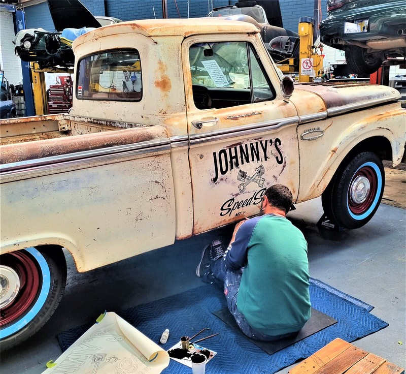 An auto mechanic in the TGS Muscle Cars shop adds a custom decal to an old style truck that bears the logo for "Johnny's Sports".