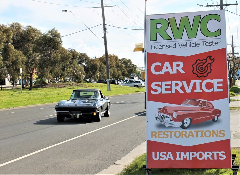 A classic car drives past a sign that designates that TGS MUscle Cars is a Roadworthy Certificate Licensed Vehicle Tester.